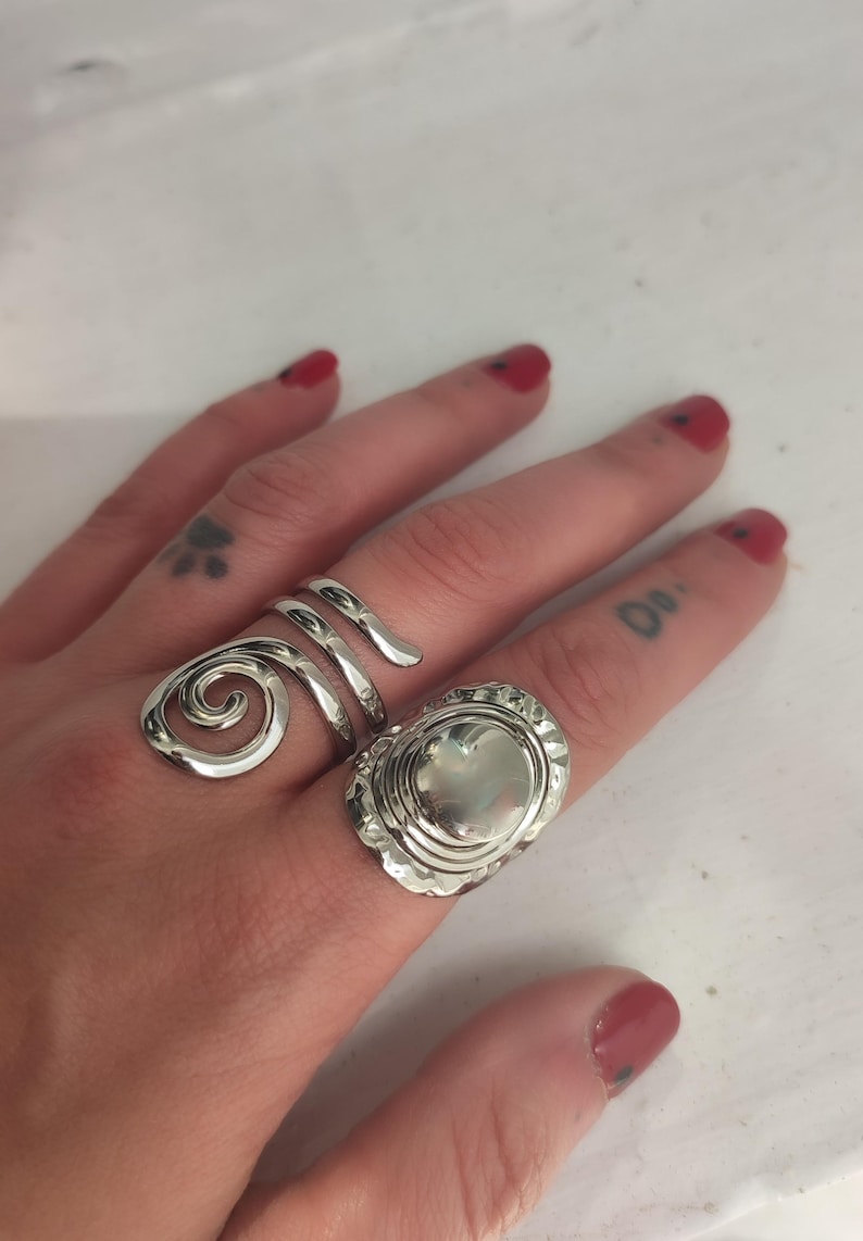 May include: Two silver rings on a finger. One ring is a spiral design and the other is a round ring with a textured surface.