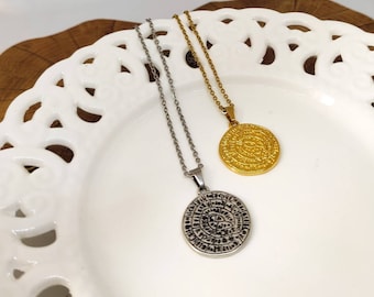 Peut inclure: Deux colliers avec des pendentifs circulaires. L'un est argenté, l'autre doré. Les deux ont une chaîne et un pendentif avec un motif gravé. Les colliers sont présentés sur une assiette blanche décorative.