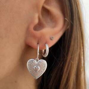Puede incluir: Pendientes plateados en forma de coraz&oacute;n con intrincados patrones florales y un coraz&oacute;n m&aacute;s peque&ntilde;o en el centro. Los pendientes cuelgan de aros plateados. La imagen muestra un primer plano de los pendientes y la oreja.
