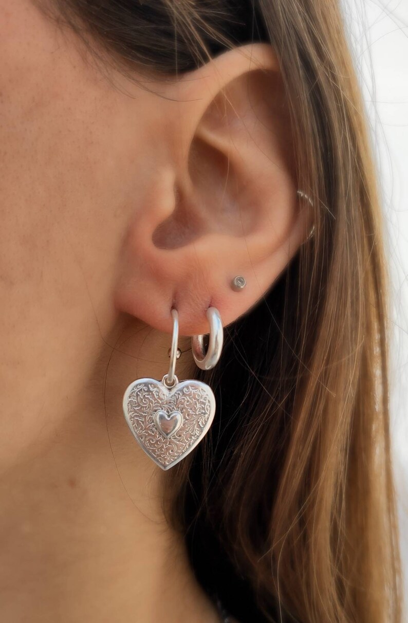 Puede incluir: Pendientes plateados en forma de coraz&oacute;n con intrincados patrones florales y un coraz&oacute;n m&aacute;s peque&ntilde;o en el centro. Los pendientes cuelgan de aros plateados. La imagen muestra un primer plano de los pendientes y la oreja.