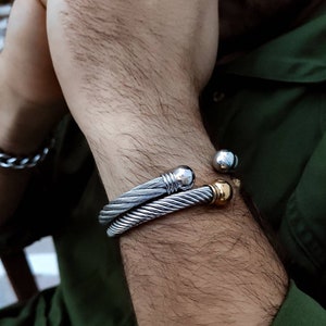K&ouml;nnte beinhalten: Zwei silberfarbene Kabelarmb&auml;nder und ein silbernes Kettenarmband sind an einem Arm zu sehen. Ein Armband hat eine silberne Kugel und einen goldfarbenen Akzent. Das andere Armband hat einen silbernen Kugelakzent.
