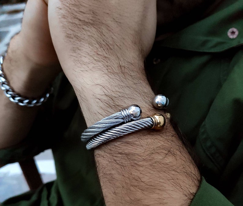 K&ouml;nnte beinhalten: Zwei silberfarbene Kabelarmb&auml;nder und ein silbernes Kettenarmband sind an einem Arm zu sehen. Ein Armband hat eine silberne Kugel und einen goldfarbenen Akzent. Das andere Armband hat einen silbernen Kugelakzent.