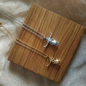 May include: Two necklaces with bull skull pendants. One necklace is silver-colored, and the other is gold-colored. The pendants are detailed with horns and facial features. The necklaces are displayed on a wooden surface.