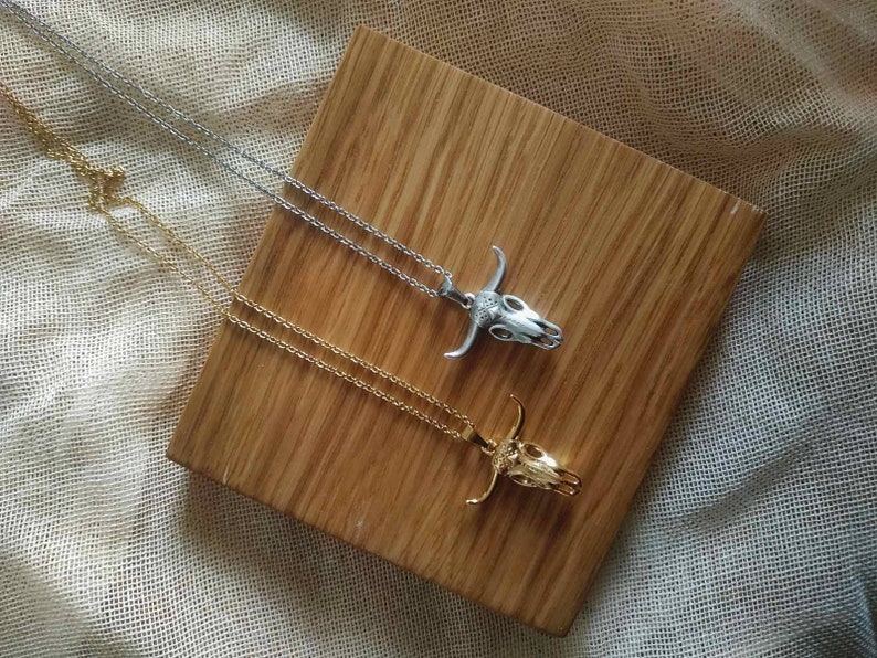 May include: Two necklaces with bull skull pendants. One necklace is silver-colored, and the other is gold-colored. The pendants are detailed with horns and facial features. The necklaces are displayed on a wooden surface.
