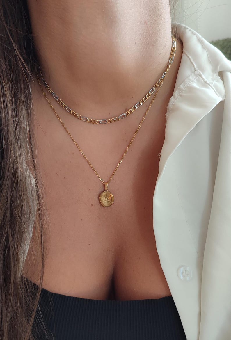 May include: A layered necklace set featuring a gold chain and a pendant necklace. The top chain is a gold and silver curb chain. The pendant is a small, round gold charm with a sunburst design. The necklaces are worn with a white button-down shirt.