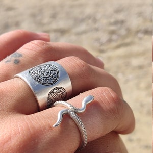 May include: Two silver rings are displayed. One is a wide band with an embossed circular design. The other is a snake ring, with the snake's head and body wrapping around the finger. The rings are on a hand, with a beach in the background.