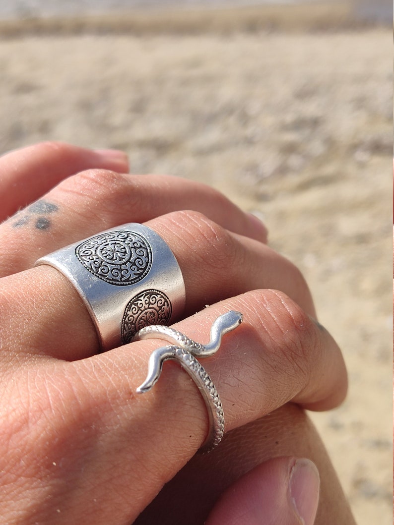 May include: Two silver rings are displayed. One is a wide band with an embossed circular design. The other is a snake ring, with the snake's head and body wrapping around the finger. The rings are on a hand, with a beach in the background.