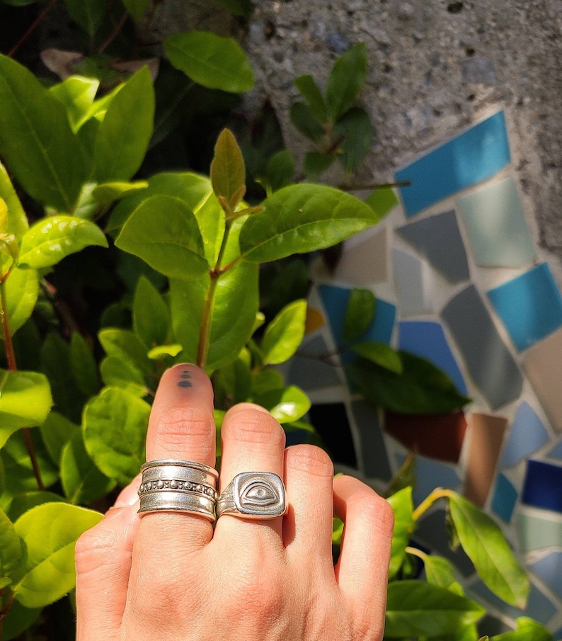 Pu&ograve; includere: Un anello d'argento con un designo a forma di occhio su una base quadrata, indossato su un dito con altri due anelli d'argento.