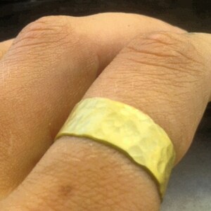 May include: A hammered gold-colored ring with a textured surface. The ring is wide and wraps around a finger. The ring's surface reflects light, creating highlights and shadows.