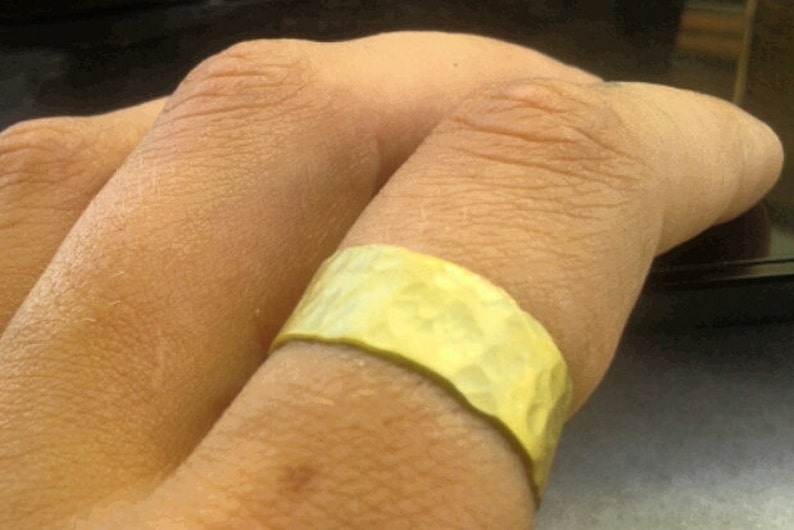 May include: A hammered gold-colored ring with a textured surface. The ring is wide and wraps around a finger. The ring's surface reflects light, creating highlights and shadows.