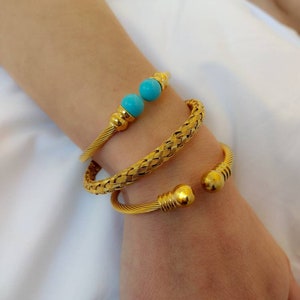 Könnte beinhalten: Goldfarbene Armbänder. Eines hat ein geflochtenes Design, das andere ein gedrehtes Kabeldesign mit türkisfarbenen Perlen und goldenen Akzenten. Die Armbänder werden am Handgelenk getragen und zeigen ihr elegantes und stilvolles Aussehen.