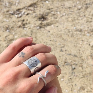 May include: Two silver rings. One is a wide band with a circular pattern. The other is a snake ring with a curved design. The rings are on a hand, with a beach background.