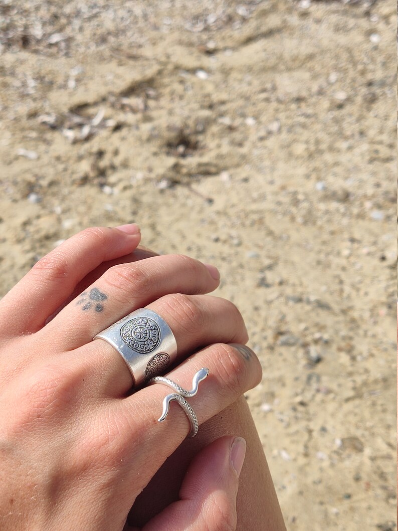 May include: Two silver rings. One is a wide band with a circular pattern. The other is a snake ring with a curved design. The rings are on a hand, with a beach background.
