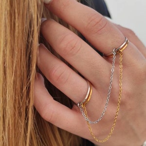 May include: Two-finger ring with a gold and silver chain. The ring has a gold band and a silver band. The chain connects the two rings. The rings are on a person's finger.