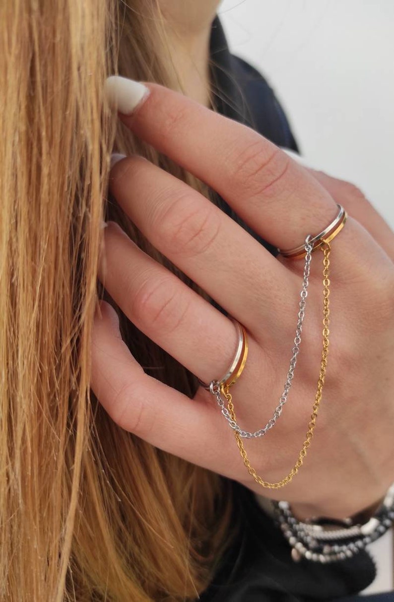 May include: Two-finger ring with a gold and silver chain. The ring has a gold band and a silver band. The chain connects the two rings. The rings are on a person's finger.
