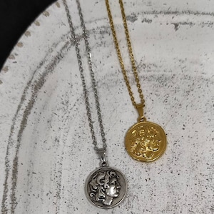Puede incluir: Dos collares con colgantes de monedas. Uno es plateado, el otro dorado. Cada collar presenta un colgante circular con el perfil de una persona. Los colgantes cuelgan de delicadas cadenas. Los collares se exhiben sobre una superficie blanca texturizada.