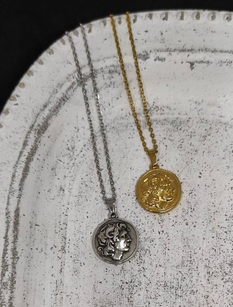 Puede incluir: Dos collares con colgantes de monedas. Uno es plateado, el otro dorado. Cada collar presenta un colgante circular con el perfil de una persona. Los colgantes cuelgan de delicadas cadenas. Los collares se exhiben sobre una superficie blanca texturizada.
