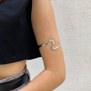 May include: A silver-toned arm cuff with a wavy design. The cuff is worn on the upper arm and has a smooth, polished finish. The jewelry is a modern accessory.