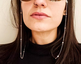 May include: A close-up shot of a person wearing sunglasses with a delicate chain eyeglass holder. The chain is silver-toned with small, round, pink and brown beads. The person is wearing a black top.