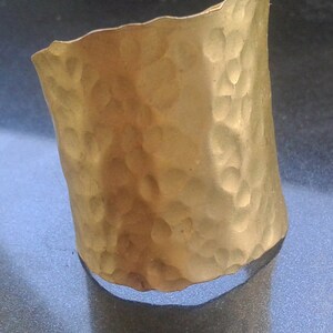 May include: A hammered gold-tone cuff bracelet. The cuff has a textured surface with a series of small, irregular indentations. The edges of the cuff are slightly uneven, giving it a handcrafted look.
