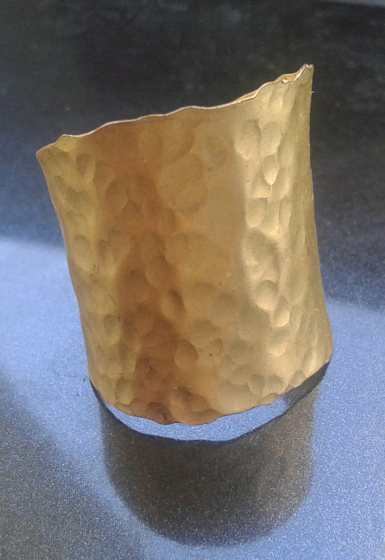 May include: A hammered gold-tone cuff bracelet. The cuff has a textured surface with a series of small, irregular indentations. The edges of the cuff are slightly uneven, giving it a handcrafted look.