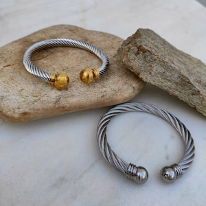 K&ouml;nnte beinhalten: Zwei silber- und goldfarbene Kabelarmb&auml;nder. Ein Armband hat goldfarbene Kugelenden. Das andere Armband hat silberfarbene Kugelenden. Die Armb&auml;nder werden auf einer hellen Steinoberfl&auml;che pr&auml;sentiert.