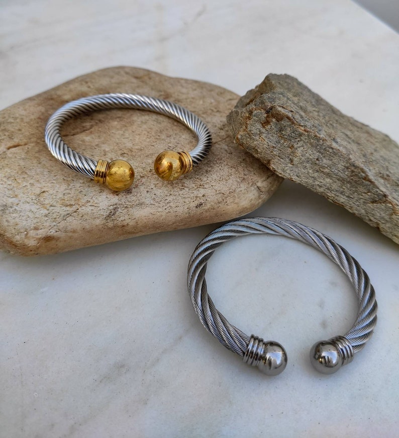 K&ouml;nnte beinhalten: Zwei silber- und goldfarbene Kabelarmb&auml;nder. Ein Armband hat goldfarbene Kugelenden. Das andere Armband hat silberfarbene Kugelenden. Die Armb&auml;nder werden auf einer hellen Steinoberfl&auml;che pr&auml;sentiert.