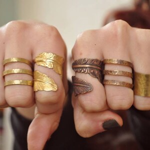 May include: A display of various metal rings. The rings include gold-toned bands, hammered bands, and feather-shaped designs. The rings are displayed on two fists, showcasing the different styles and textures of the jewelry.