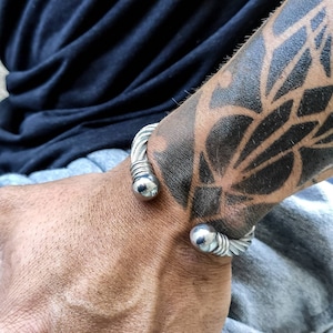 K&ouml;nnte beinhalten: Ein silberfarbenes Armreif-Armband mit einem geflochtenen Kabeldesign und kugelf&ouml;rmigen Endkappen. Das Armband wird an einem Handgelenk mit einem gro&szlig;en schwarzen Tattoo getragen.