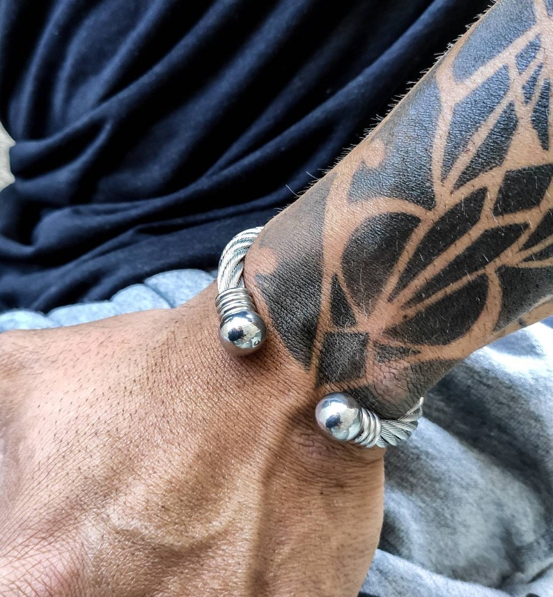 K&ouml;nnte beinhalten: Ein silberfarbenes Armreif-Armband mit einem geflochtenen Kabeldesign und kugelf&ouml;rmigen Endkappen. Das Armband wird an einem Handgelenk mit einem gro&szlig;en schwarzen Tattoo getragen.