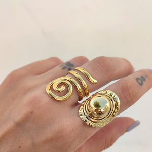 May include: Two gold rings. One has a spiral design, the other an oval face with a raised centre. Both rings are worn on a hand. The rings are made of a shiny metal.