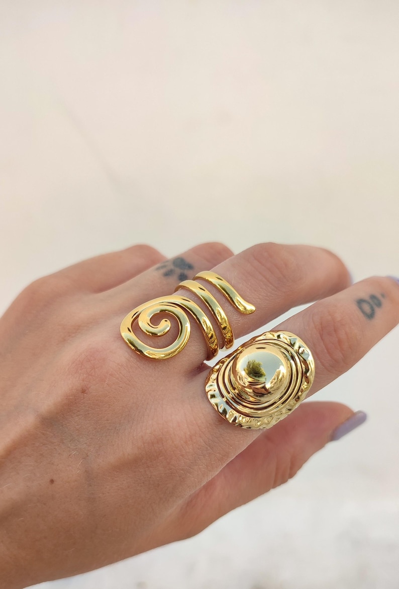 May include: Two gold rings. One has a spiral design, the other an oval face with a raised centre. Both rings are worn on a hand. The rings are made of a shiny metal.