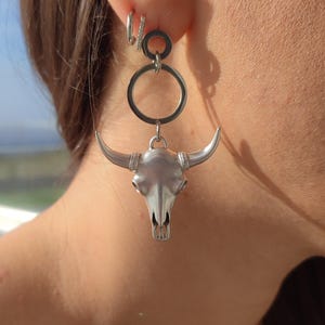 K&ouml;nnte beinhalten: Ohrringe mit silberfarbenem Bullensch&auml;del. Die Ohrringe haben einen detaillierten Bullensch&auml;del-Anh&auml;nger, der an einem kreisf&ouml;rmigen Ring und einem kleineren Ring befestigt ist. Das Design ist ein auff&auml;lliges Statement-St&uuml;ck, perfekt f&uuml;r einen Hauch von Western-Flair.