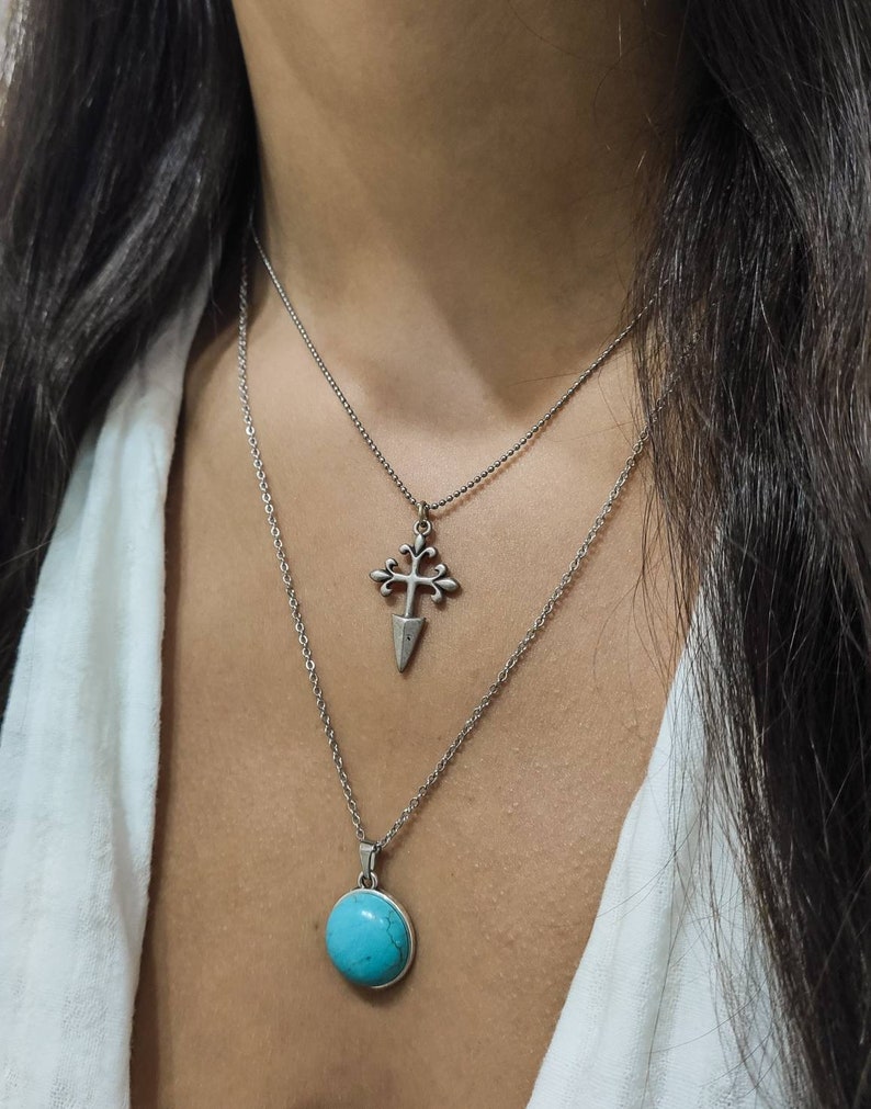 May include: A layered necklace featuring two pendants. The top pendant is a silver cross design, and the bottom pendant is a round, turquoise stone. Both pendants hang from delicate silver chains, creating a stylish accessory.