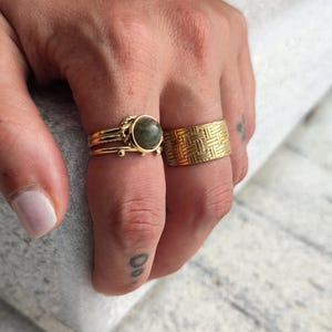 Op de afbeelding: Twee gouden ringen aan een vinger. De ring aan de wijsvinger heeft een groene steen. De ring aan de middelvinger heeft een geometrisch patroon.