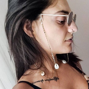 May include: Gold-toned eyeglass chain with a delicate chain design and seashell accents. The chain is attached to the glasses with clear rubber loops. The chain is worn around the neck.