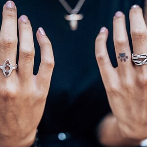 Pu&ograve; includere: Una collezione di cinque anelli in argento indossati sulle dita. Gli anelli presentano vari design, tra cui una barra verticale, un intaglio a forma di occhio, un motivo a corda annodata e un anello a sigillo quadrato con un occhio inciso.