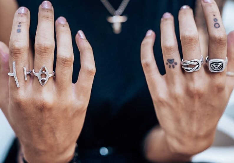 Pu&ograve; includere: Una collezione di cinque anelli in argento indossati sulle dita. Gli anelli presentano vari design, tra cui una barra verticale, un intaglio a forma di occhio, un motivo a corda annodata e un anello a sigillo quadrato con un occhio inciso.