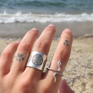 May include: Two silver rings on a hand, one a wide band with an embossed circular design, the other a snake ring. The snake ring has a coiled body and a raised head. The background is a beach with ocean waves.