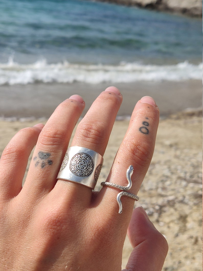 May include: Two silver rings on a hand, one a wide band with an embossed circular design, the other a snake ring. The snake ring has a coiled body and a raised head. The background is a beach with ocean waves.