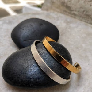 Pode incluir: Duas pulseiras minimalistas, uma dourada e outra prateada, repousam sobre uma pedra preta lisa. A pulseira dourada tem um acabamento polido e quente, enquanto a pulseira prateada tem um aspeto mate e frio. As pulseiras s&atilde;o simples e elegantes.