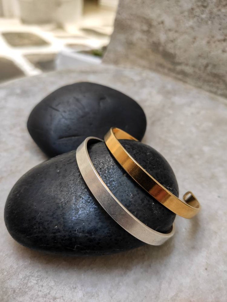 Pode incluir: Duas pulseiras minimalistas, uma dourada e outra prateada, repousam sobre uma pedra preta lisa. A pulseira dourada tem um acabamento polido e quente, enquanto a pulseira prateada tem um aspeto mate e frio. As pulseiras s&atilde;o simples e elegantes.