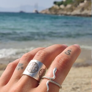 May include: Two silver rings on a finger. One is a wide band with a circular pattern. The other is a snake ring with a coiled body and a raised head. The background is a beach with blue water and a clear sky.