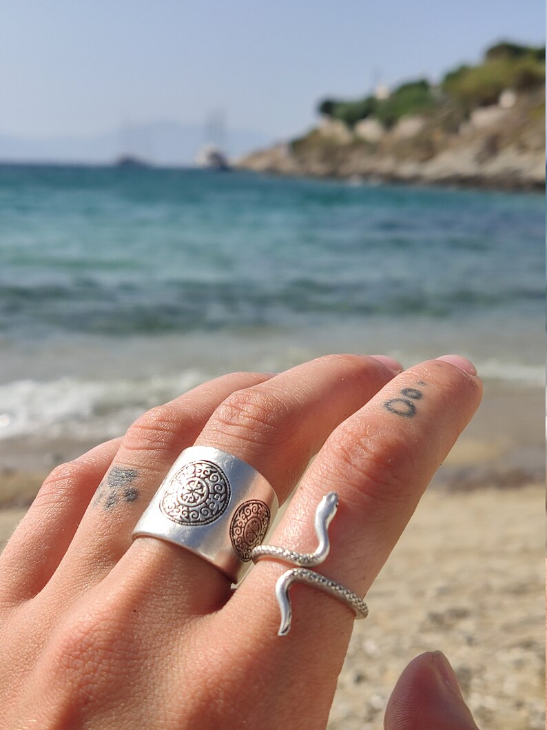 May include: Two silver rings on a finger. One is a wide band with a circular pattern. The other is a snake ring with a coiled body and a raised head. The background is a beach with blue water and a clear sky.