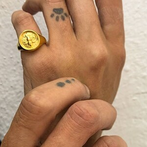 Op de afbeelding: Er worden twee ringen getoond, &eacute;&eacute;n goud en &eacute;&eacute;n zilver. De gouden ring heeft een gedetailleerd bijenontwerp, terwijl de zilveren ring een vergelijkbaar bijenmotief heeft. De ringen worden aan vingers gedragen, met een pootafdruk-tattoo zichtbaar.