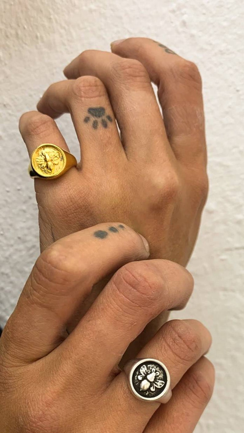 Op de afbeelding: Er worden twee ringen getoond, &eacute;&eacute;n goud en &eacute;&eacute;n zilver. De gouden ring heeft een gedetailleerd bijenontwerp, terwijl de zilveren ring een vergelijkbaar bijenmotief heeft. De ringen worden aan vingers gedragen, met een pootafdruk-tattoo zichtbaar.
