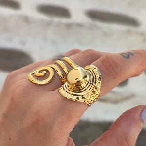 May include: A gold-coloured statement ring with a textured design and a central, raised circular element. The ring has a unique, swirling accent. The ring is worn on a finger.