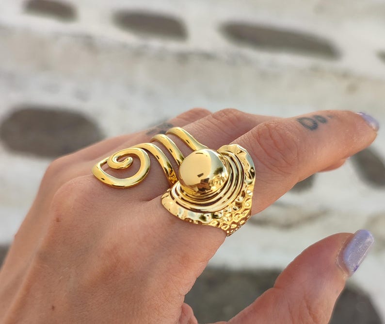 May include: A gold-coloured statement ring with a textured design and a central, raised circular element. The ring has a unique, swirling accent. The ring is worn on a finger.