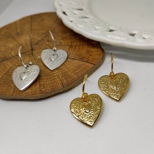 Puede incluir: Pendientes plateados y dorados en forma de coraz&oacute;n. Los pendientes tienen un patr&oacute;n floral detallado y un coraz&oacute;n m&aacute;s peque&ntilde;o en el centro. Los pendientes est&aacute;n sobre una superficie de madera.