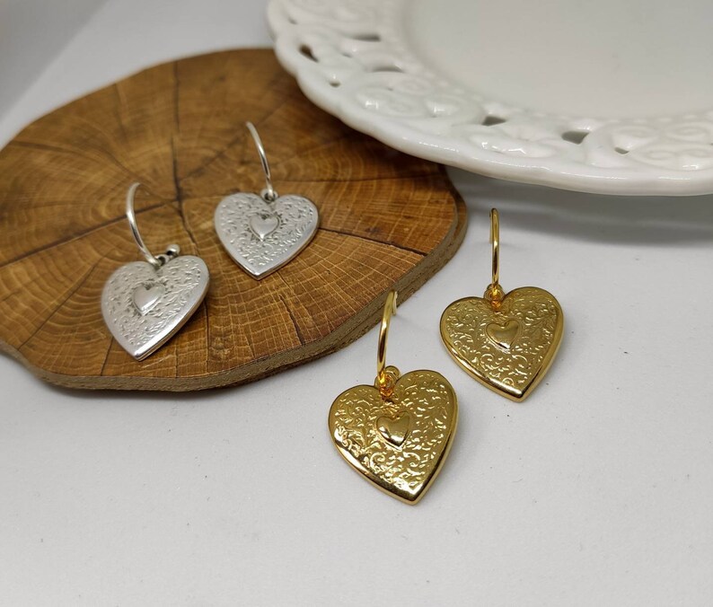 Puede incluir: Pendientes plateados y dorados en forma de coraz&oacute;n. Los pendientes tienen un patr&oacute;n floral detallado y un coraz&oacute;n m&aacute;s peque&ntilde;o en el centro. Los pendientes est&aacute;n sobre una superficie de madera.